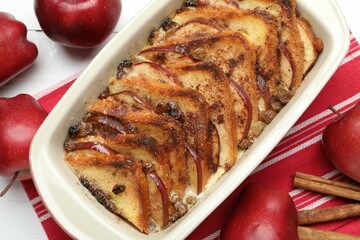 Delicious bread pudding in baking dish, fresh apples and cinnamon sticks on white table, flat lay