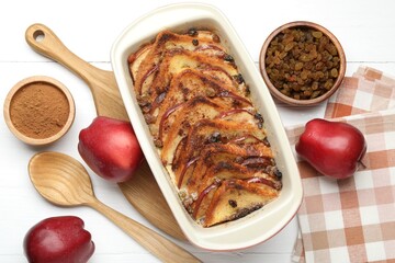 Delicious bread pudding in baking dish, fresh apples, cinnamon and raisins on white wooden table, flat lay
