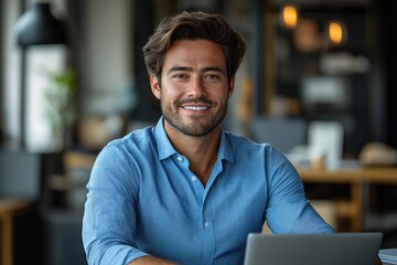 Fototapeta premium Young confident man smiles at camera working on laptop in office. Man sits at desk, looks happy. Modern businessman in blue shirt using tech. Success in business occupation. Concentrated on project.