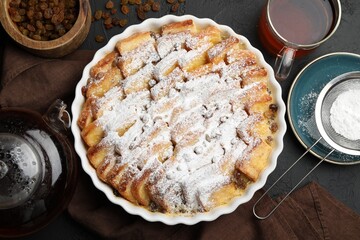 Delicious bread pudding with raisins, powdered sugar and tea on black table, flat lay