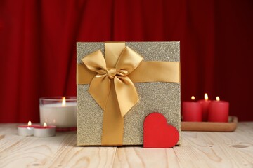 Beautiful gift box, decorative heart and burning candles on wooden table against red curtain....