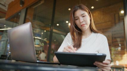woman using tablet while sitting at table with laptop nearby, focused on her work in modern cafe. businesswoman remote work digital nomad