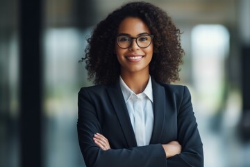 Portrait of a smiling businesswoman wearing glasses and a suit with arms crossed, exuding confidence and professionalism in a modern office environment