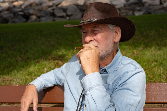 Portrait of an old smiling caucasian senior bearded man in outdoors wearing a cowboy leather hat smoking a cigarillo