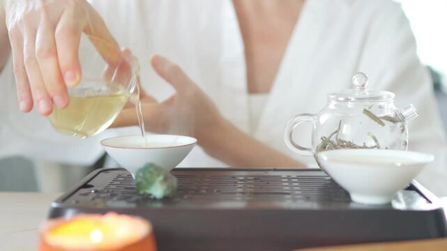 Female pouring tea from clear glass pitcher teaware into tea cups, creating serene tea ritual. Soft light, wooden tea tray evoke calming mindful Chenese ceremony atmosphere 4K video