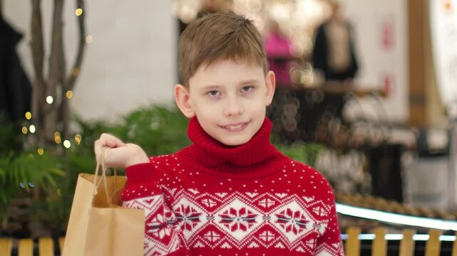 Portrait of cute Caucasian boy 10 years old in red New Year sweater with shopping bag while in shopping center looking at camera