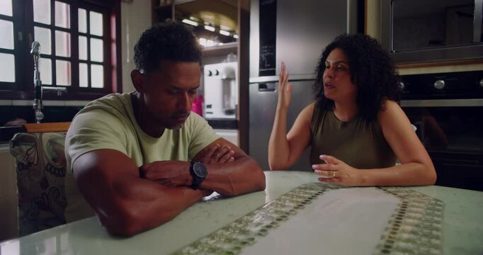 Husband visibly annoyed and frustrated while wife discusses concerns at dining table, unresolved relationship dynamics in modern kitchen