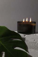 Burning soy wax candle on concrete block and monstera leaf against gray background, space for text