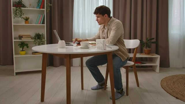 Young man in the morning in the kitchen dinning room enjoying healthy breakfast, sitting at table finishes breakfast and work on laptop, takes notebook and walks away.