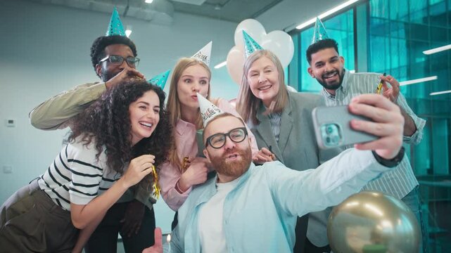 Handsome male sitting at his desk at work while celebrating birthday together with colleagues. Holding smartphone with one hand and making selfie or recording video. Blowing out candles.
