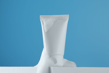 Tube of cosmetic product with foam on podium against light blue background