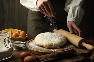 Woman with rolling pin adding flour onto dough at wooden table, closeup