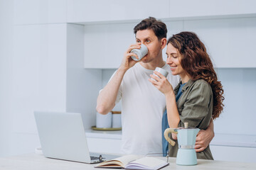 Young couple embracing and sipping coffee while using a laptop in a modern kitchen, sharing a cozy morning routine filled with love and connection in their bright, airy home
