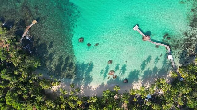 Aerial view of a tropical beach with turquoise waters, palm trees, and a wooden pier extending into the sea, showcasing natural beauty and a serene environment