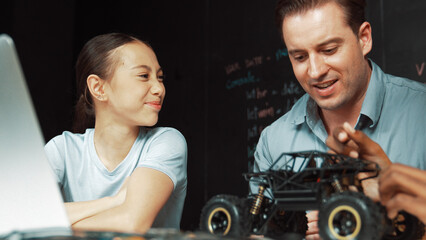 Smart teacher inspect car model construction to caucasian girl. Highschool student with mixed races listening mentor explain robotic model system at table with laptop and wires placed. Edification.