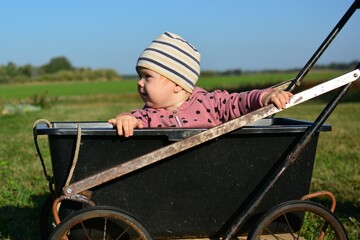 Baby girl is sitting in hand made wheelbarrow in rural area. Cute toddler girl sitting in old baby stroller in countryside in sunny day. Poor happy childhood.