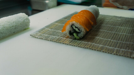 The chef prepares a Philadelphia sushi roll with fresh salmon slices on top and creamy cheese inside, placed on a white cutting board. Wearing disposable gloves, the process is captured in a close-up 