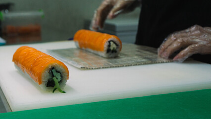 The chef prepares a Philadelphia sushi roll with vibrant red fish on top and creamy cheese filling, carefully arranged on a white cutting board while wearing disposable gloves. A close-up view