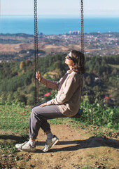 Woman sitting on a wooden swing on a hilltop, enjoying the serene view of the sea and countryside...