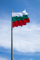 The Bulgarian flag, waving on a pole with a blue sky with white clouds in the background.