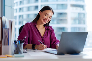 Happy European business woman CEO working on laptop and taking notes, sitting at desk in office....