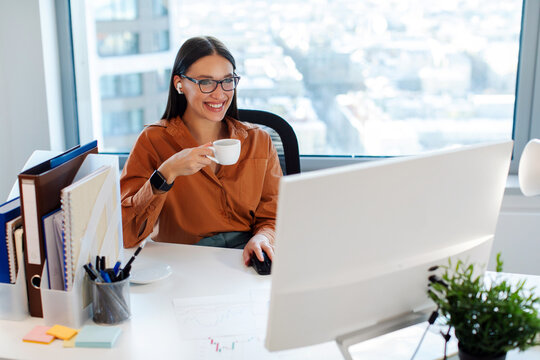 Good day at work. Happy European businesswoman drinking coffee at workplace, looking at computer monitor screen and smiling, free space