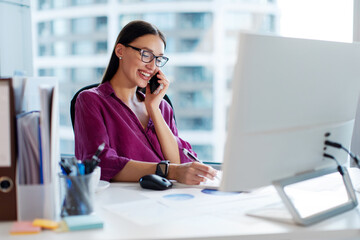Female manager talking on cellphone and taking notes, sitting at her desk in office, working at...