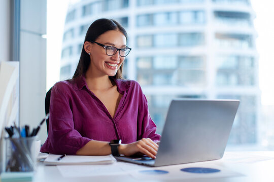 Young happy woman office worker employee sitting at desk working on laptop, doing project, checking emails or chatting with clients online - Powered by Adobe