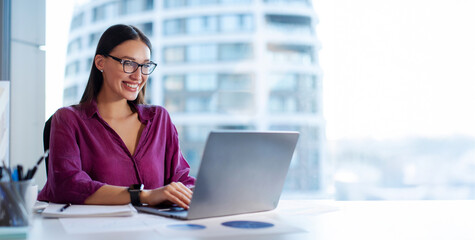 Successful young woman working on laptop in office, tech support worker typing on keyboard smiling, sitting at desk, panorama with free space