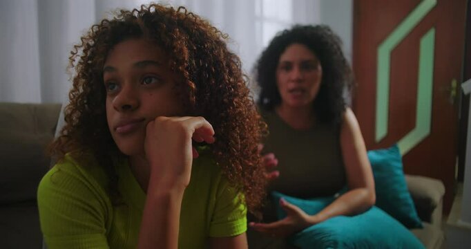 Teenager daughter ignoring mother, sitting on couch with head resting on hand, looking away with disinterest, mother covering face in frustration in family living room
