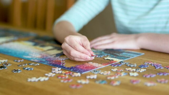 Close-up view of young woman hands assembling puzzles at home. Connecting jigsaw puzzle pieces, assembling a jigsaw puzzle. Fun family leisure. Table games, home relaxation. Slow motion footage.