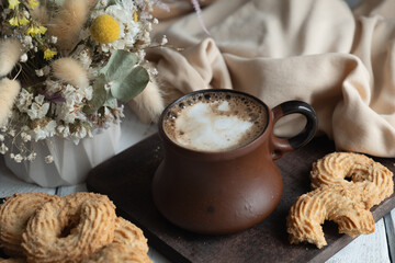 A cup of cappuccino in a ceramic brown mug on a wooden board. Nearby lie crumbly cookies in the shape of curls. The background is beige textiles and a vase of dried flowers.