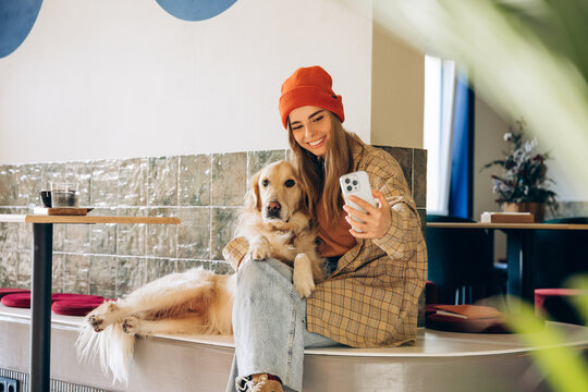 Young woman taking selfie with golden retriever in modern cafe
