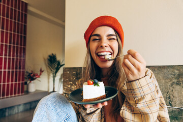 Cheerful young woman wearing orange beanie enjoying a slice of cake at a coffee shop