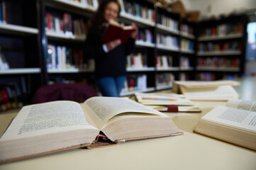 Stacks of open books lie on a table in a library as a woman reads in the background, symbolizing education, learning, and the pursuit of knowledge in an academic setting.