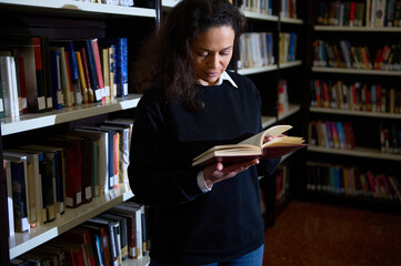 A person fully engrossed in reading a book while standing in a well-organized library, surrounded...
