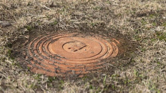 A rusty iron hatch with a pattern covered dirt above an observation well is located next to a dry grass in the daytime. Close-up