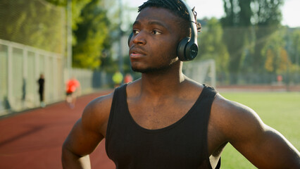 Pensive African American man sportsman athlete in headphones use smartwatch in city prepare for sport training male runner listen music on break running cardio workout outdoors morning jog on stadium