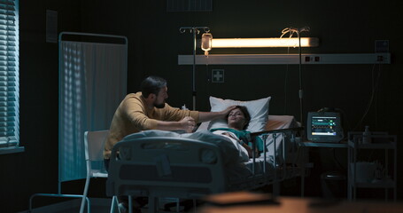 Young man sits in the hospital room beside an unconscious girl lying in the bed.