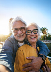 Vertical. Happy senior couple taking selfie outdoors in a park of the city. Smiling elderly people traveling together on vacation. Cheerful man and woman posing for photo with mobile phone.
