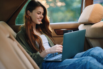 Young woman working on a laptop inside a car, smiling, wearing casual clothes, with long hair,...