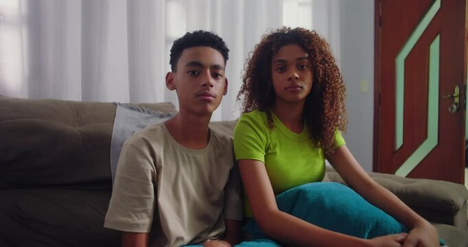 Teenager siblings sitting side by side on couch, looking directly at camera with neutral expressions, reflecting sibling presence and individuality