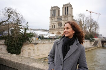 A smiling woman enjoys a breezy winter day near Notre-Dame Cathedral in Paris, France, standing by the Seine River with the historic landmark in the background.
