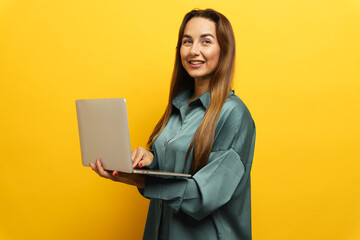 Smiling woman holding a laptop against a vibrant yellow background with casual attire