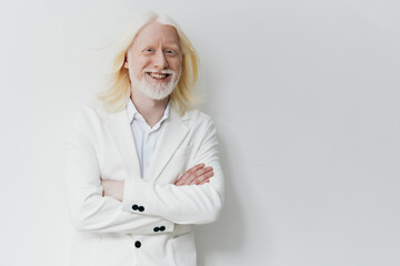 smiling man with white hair in a white suit standing with arms crossed against a light background,...