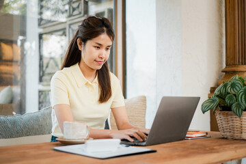 A young woman focused on her laptop at a home office desk, surrounded by coffee and plants, in a bright and cozy setting.