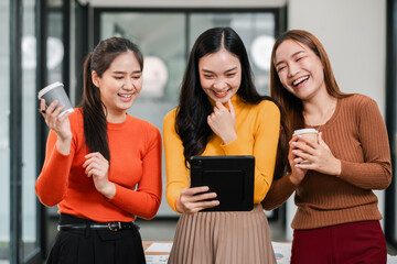 Three smiling women in vibrant sweaters share a joyful moment with coffee and a tablet in a bright, modern office.