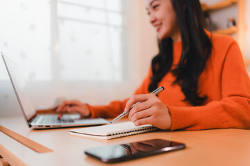 woman in orange sweater works on laptop while taking notes in cozy home office