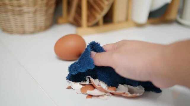 Hand cleaning up broken eggshells with a blue cloth on a white countertop, a whole brown egg and wicker basket blurred in the background