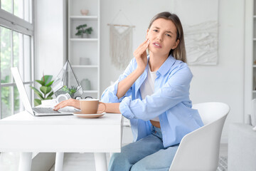 Young woman with laptop and cup of hot coffee suffering from tooth ache at home